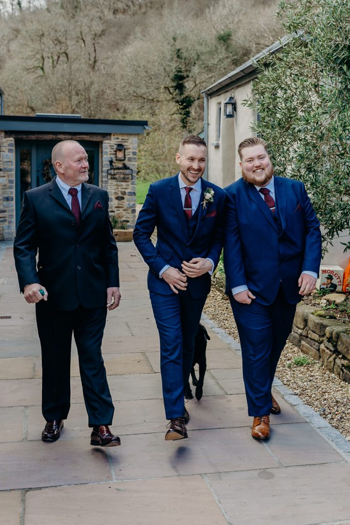 groom black dog best man and father in blue suits and burgundy ties walking down a paved path with winter woodlands and stone walls in the background