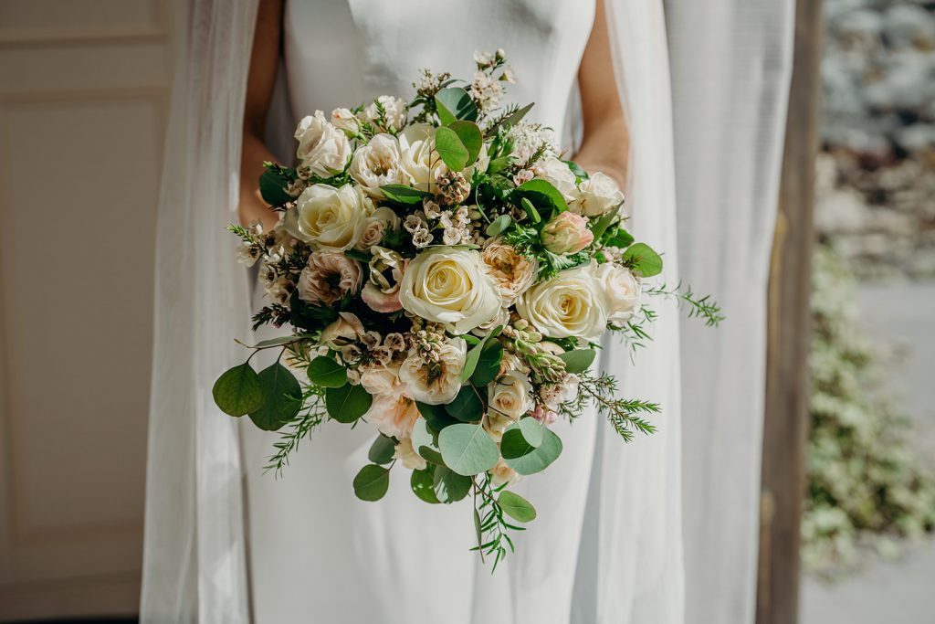 close up of an ivory and blush bridal bouquet held by bride in white dress with veil standing by an open window