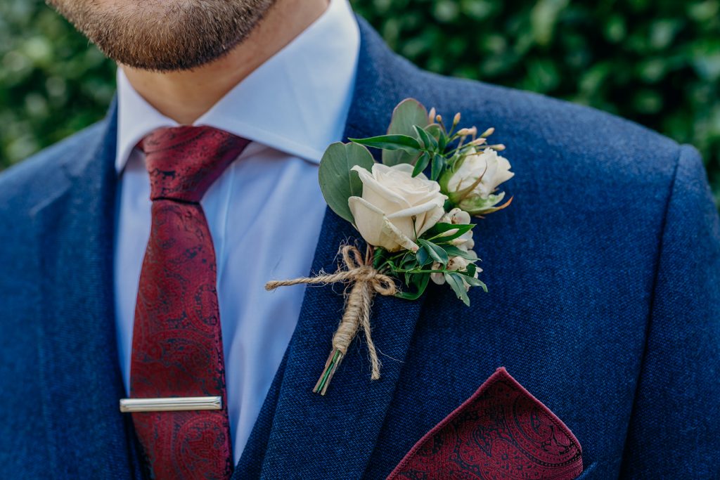 close up of a groom's white rose buttonhole with pink jasmine tied with string wearing a blue suit burgundy paisley tie & matching handkerchief square