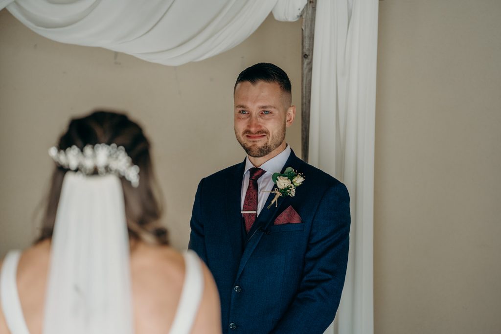 groom looking at bride with tears in his eyes as she walks into their wedding ceremony under a wooden arch draped with ivory fabric