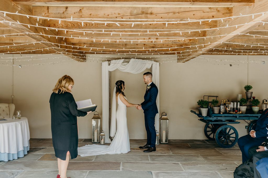 bride and groom facing each other side on holding hands getting married in a wedding barn with fairy lights stood under a wooden arch draped with ivory fabric