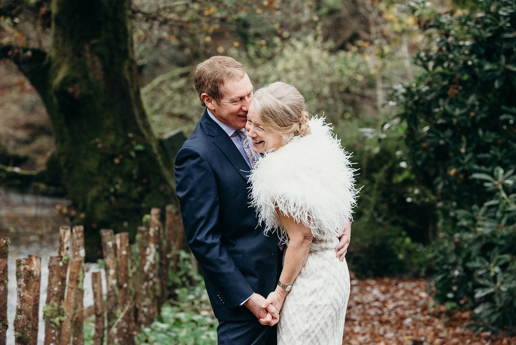 older bride and groom holding hands and laughing in a rustic outdoor setting bride wearing white feather cape 