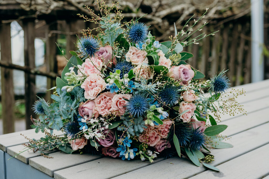 pink rose and blue thistle bridal bouquet set on a grey slatted wooden table outdoors
