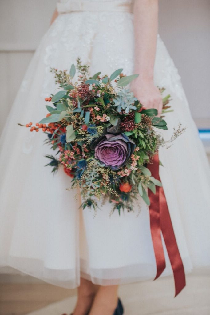 close up of bride's winter theme bouquet made up of red, purple, and navy flowers plus thistles and lots of greenery