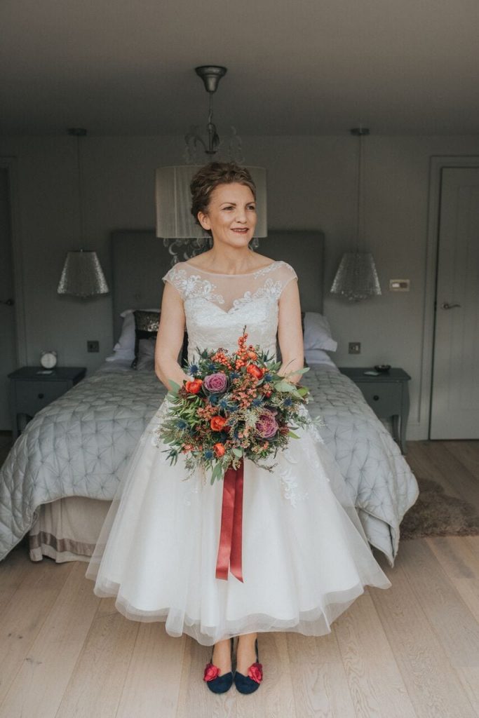 bride in her dress holding her bouquet of red, purple, and navy blue flowers with lots of greenery. her shoes are navy with a red flower and her dress is tea-length.