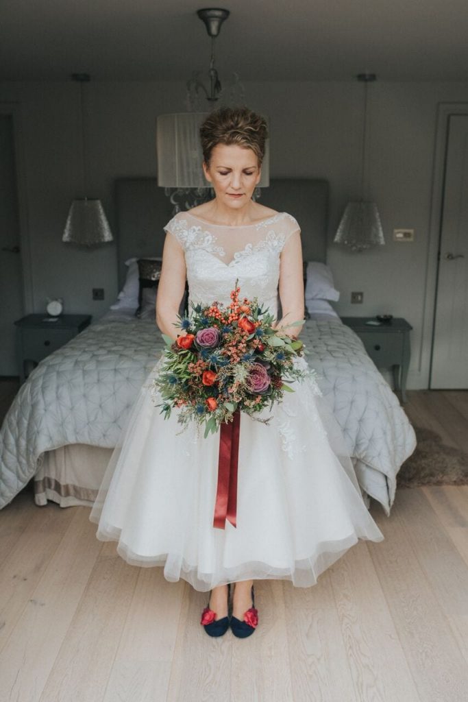 bride in honeymoon cottage holding her bright red and green bouquet. she wears a tea-length dress and has navy shoes with red flowers 