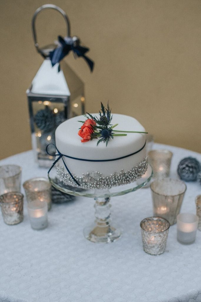 miniature elopement wedding cake with white icing, silver edible balls and the base and a navy ribbon around the middle. decorated on top with a red flower and thistles