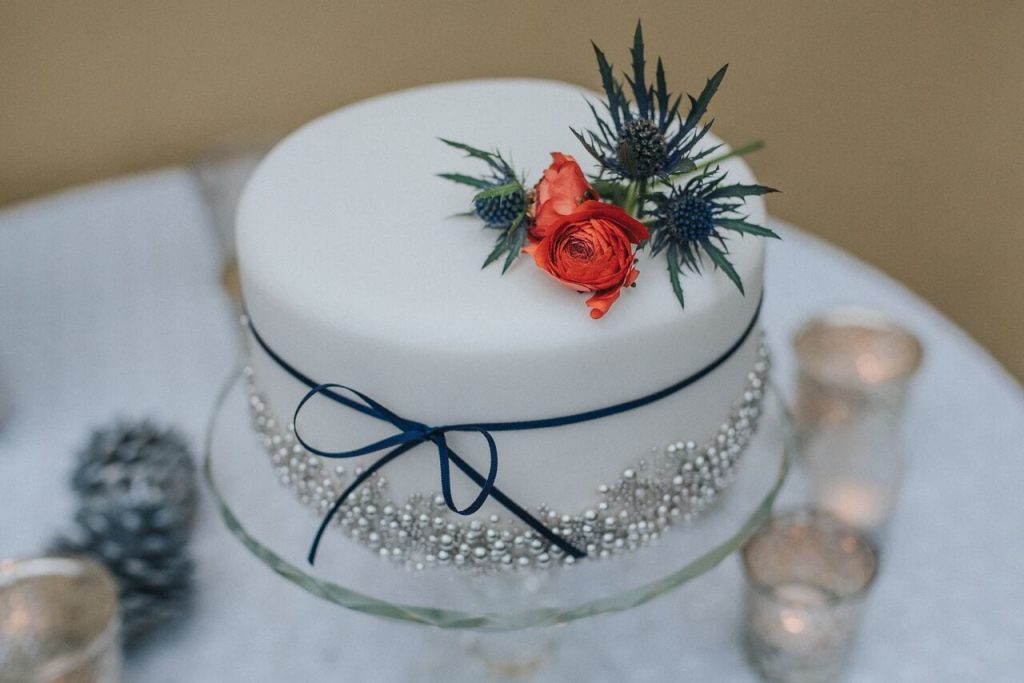 miniature elopement wedding cake with white fondant icing decorated with edible silver balls, red flowers and thistles with a thin navy ribbon around the middle.