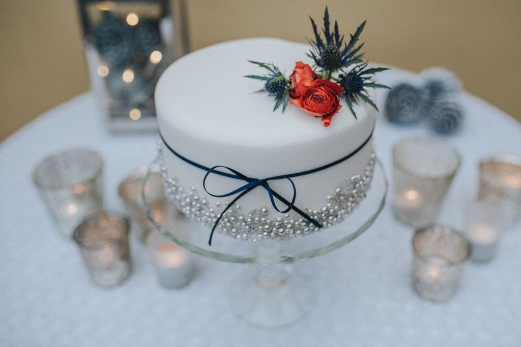 miniature elopement wedding cake with white icing, silver edible balls and the base and a navy ribbon around the middle. decorated on top with a red flower and thistles 