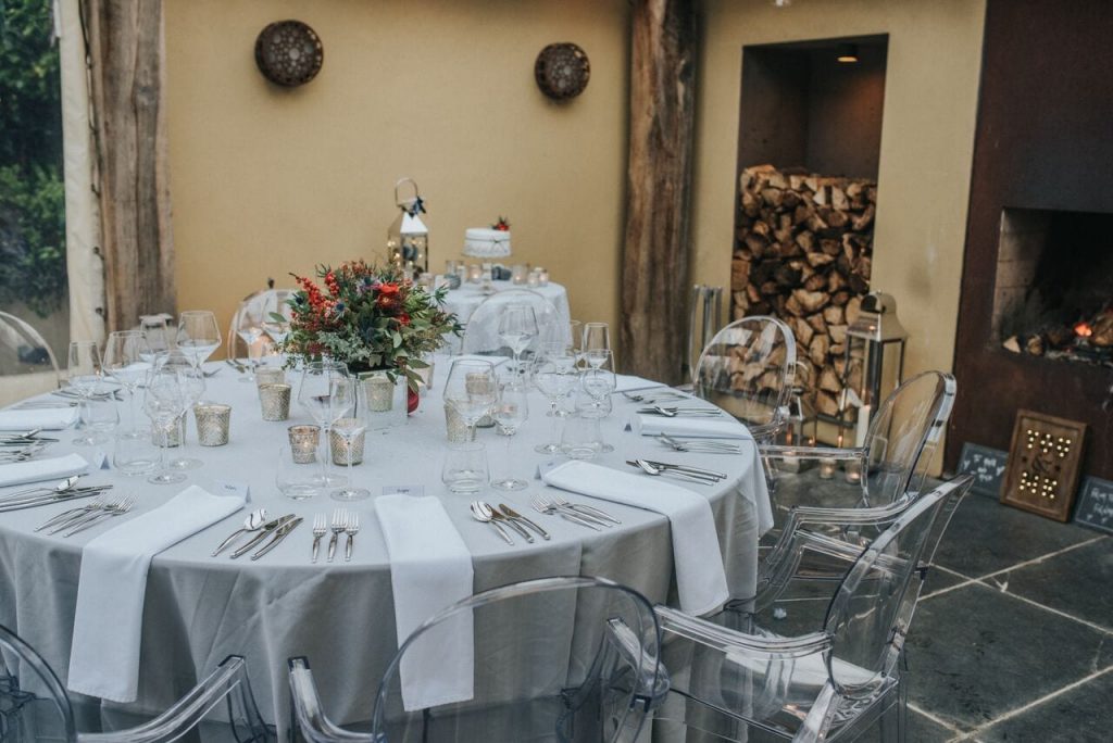 wedding breakfast table with silver grey table cloth, white napkins and ghost chairs. fire, chopped logs and cake table are in the background.