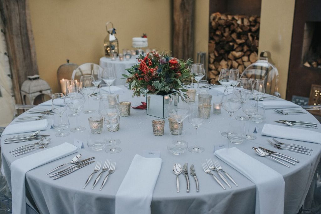 wedding breakfast table without the food. table decorated with silver grey tablecloths and white napkins with tea lights and bride's bouquet. chopped logs and cake table in the background 