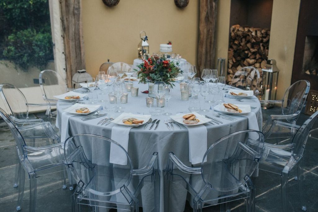 wedding breakfast table with food, silver grey tables cloths, white napkins and ghost chairs. bride's bouquet is the centrepiece and chopped logs plus their miniature cake is in the background.