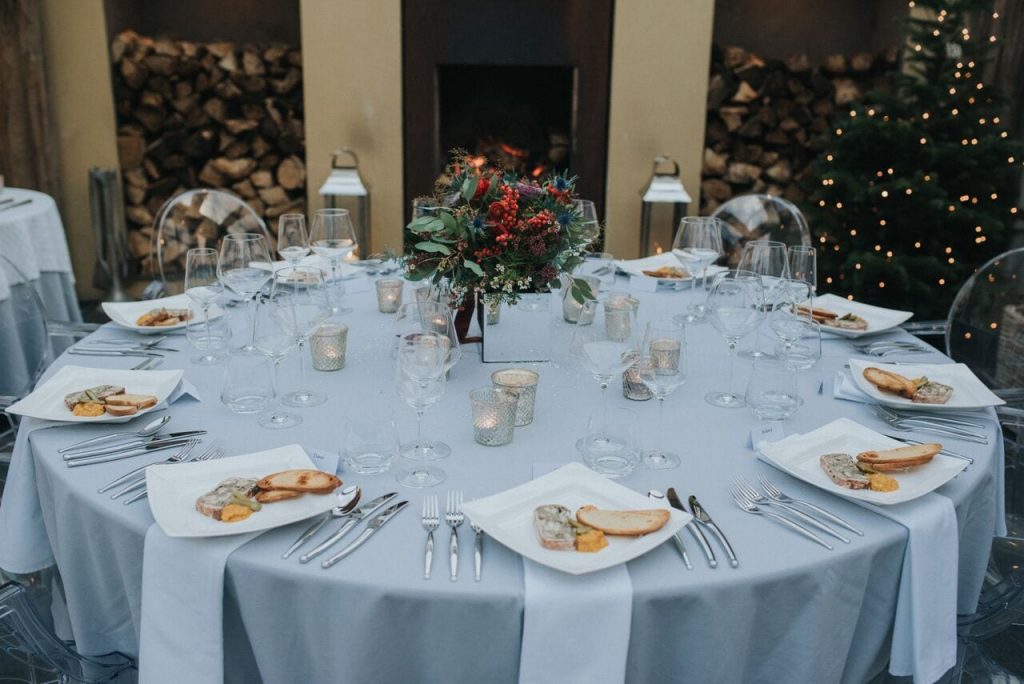 wedding breakfast table with food served on white plates. table decorated with silver grey tablecloth, white napkins, tea lights and bride's bouquet. The christmas tree and two stacks of chopped logs in the background 