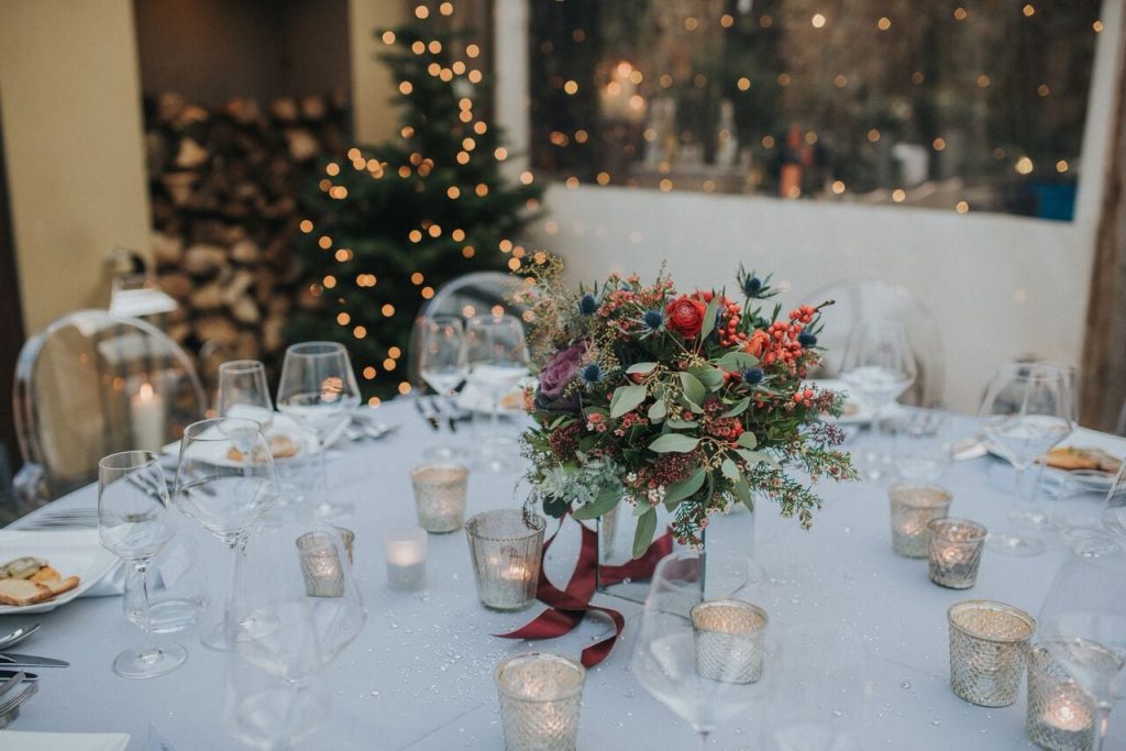 wedding breakfast table with a silver grey table cloth and tea lights, with the bride's bouquet in the centre and the christmas tree and chopped logs out of focus in the background 