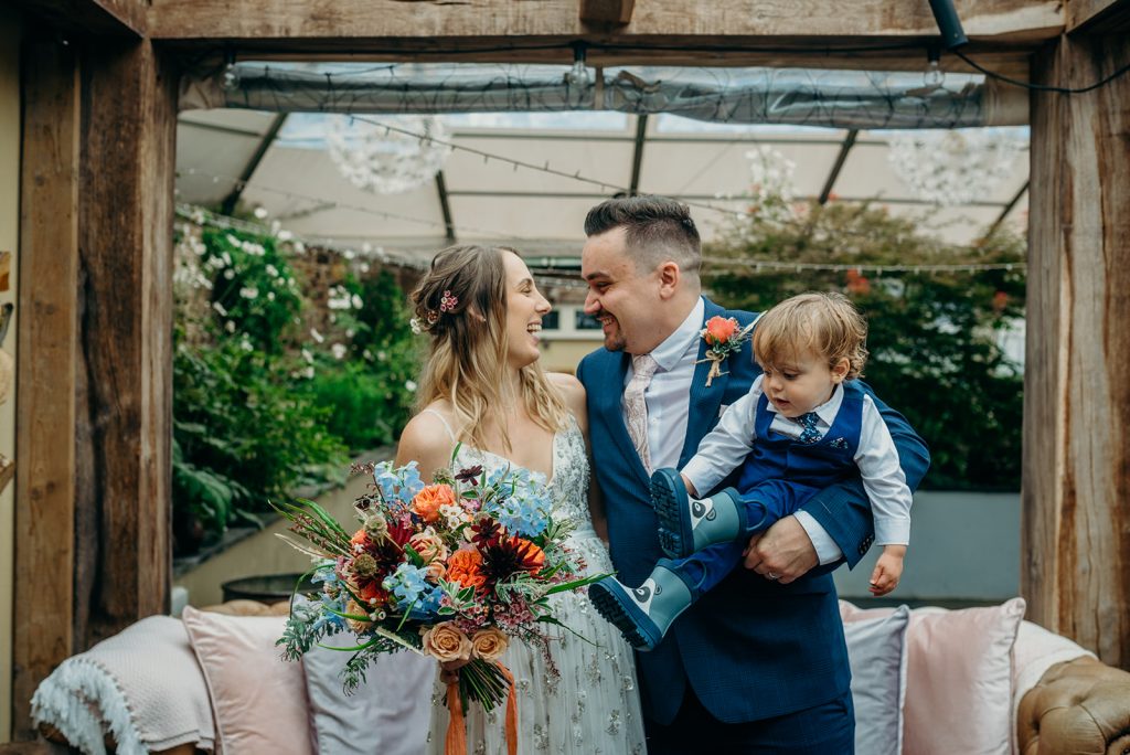 bride and groom looking at each other and smiling groom holding toddler child dressed in a suit with wellies