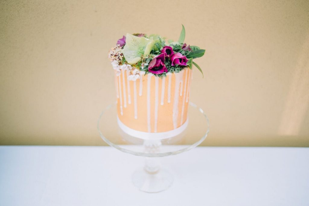 miniature elopement wedding cake with orange/gold icing and ivory icing dribbled down the sides. decorated with a large selection of deep raspberry flowers and greenery