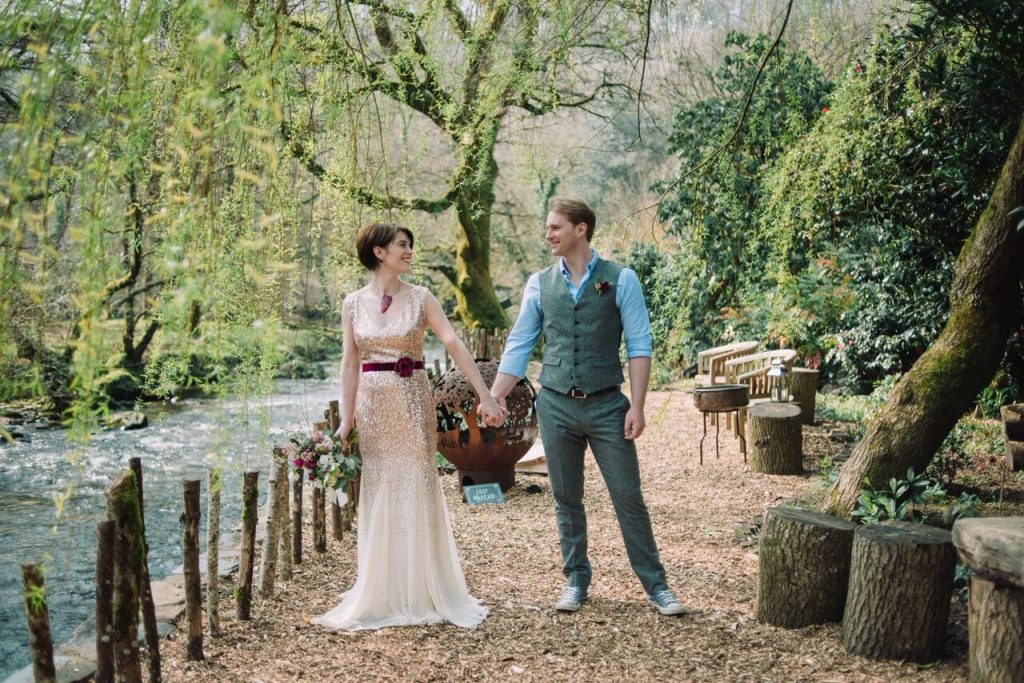 bride and groom holding hands on the riverwalk beneath the willow tree. bride wears a gold sparkly dress with maroon ribbon on waist and groom wears green tweed trousers and waistcoat with a pale blue shirt.