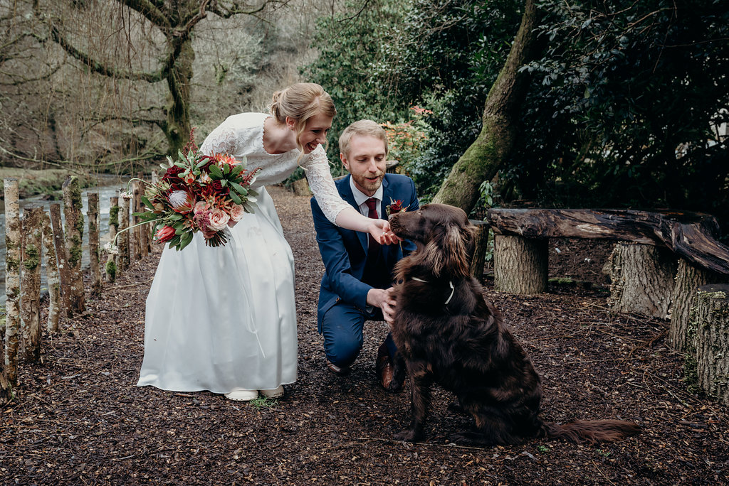 bride carrying a wedding bouquet and groom by a dartmoor river bending down to pet their Irish setter dog
