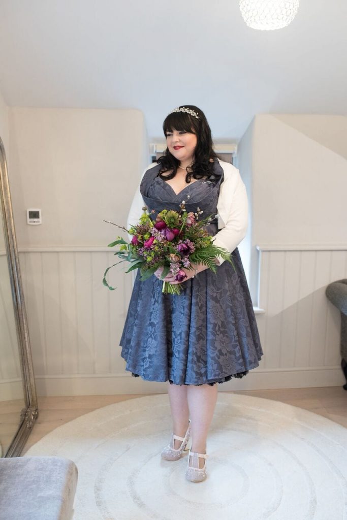 bride in tea-length dark grey dress with ivory cardigan and green and purple bouquet. 