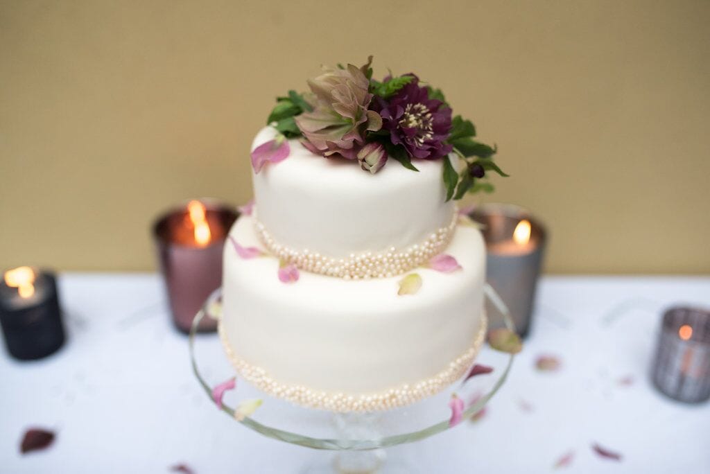 two tier miniature elopement wedding cake with ivory fondant icing decorated with cream edible balls and deep purple and green flowers and petals.