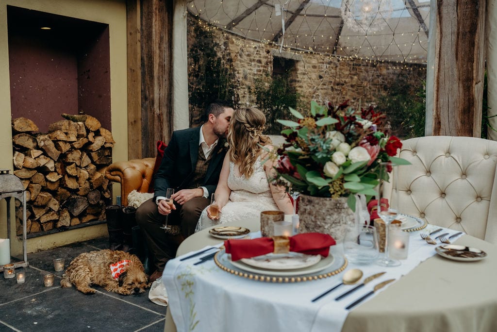 bride and groom sitting on a sofa kissing by a table laid for two with a border terrier sleeping at their feet