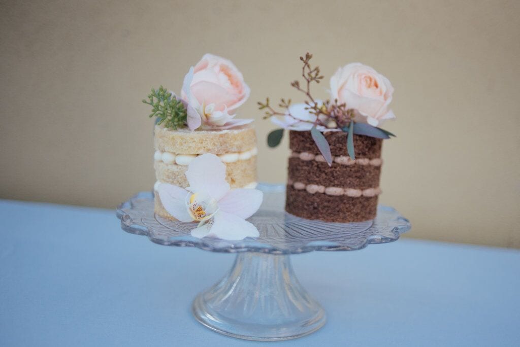 very miniature elopement wedding cakes side by side, one for the bride and one for the groom. The bride's is a naked cake with vanilla sponge and the groom's is a naked cake with chocolate sponge. both decorated with large blush pink flower and accompanying shrubbery.