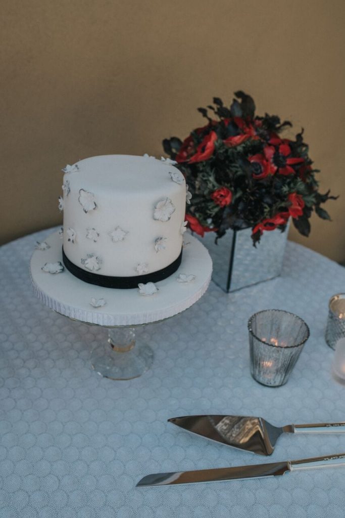 miniature elopement wedding cake with ivory fondant icing and fondant icing flowers and a black ribbon around the base to decorate. Accompanied by a vase of red flowers in the background.
