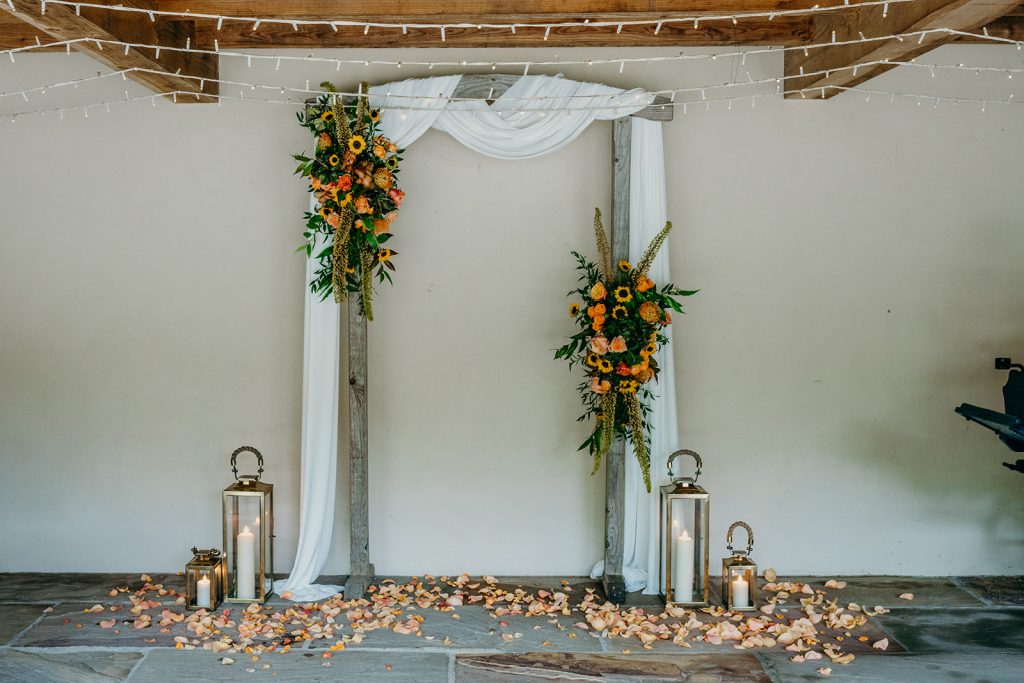 wedding ceremony backdrop with wooden arch, draped ivory fabric and 2 large flower installations in colours of apricot coral and bronze, lanterns and apricot rose petals on the floor on the floor 