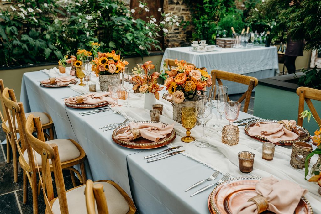 wedding tablescape with orange flowers, candles, wooden chairs and greenery backdrop
