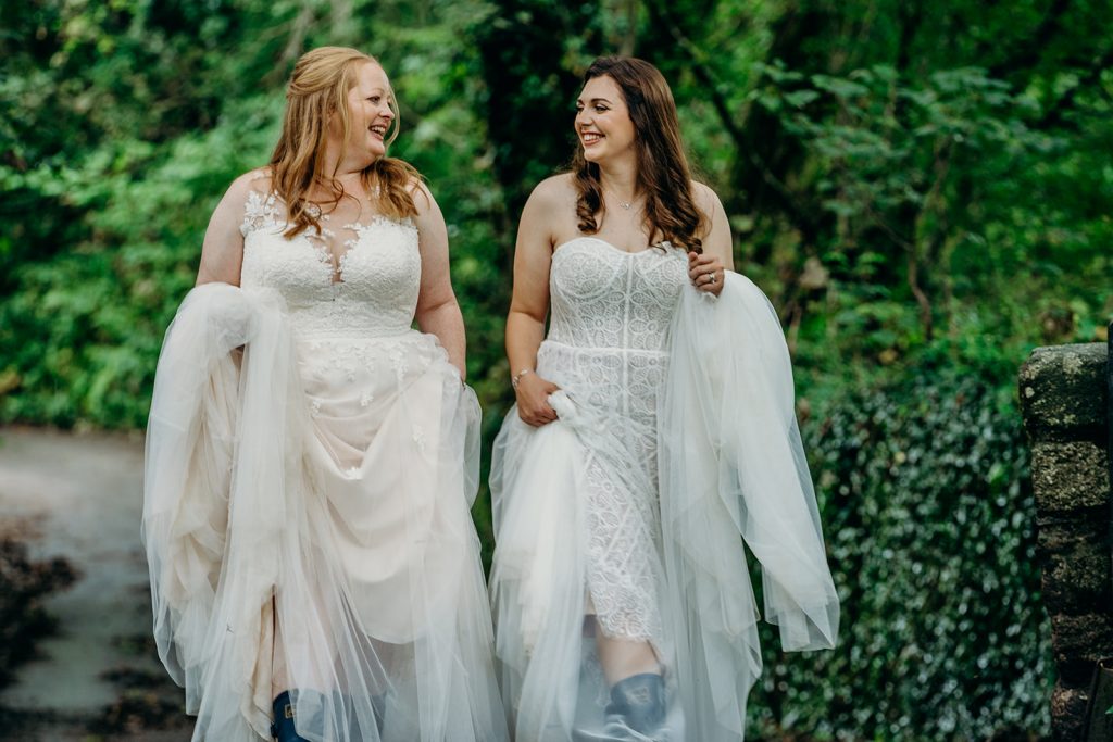 2 brides with long hair wearing white wedding dresses walking down a devon lane smiling at each other and carrying their dress skirts