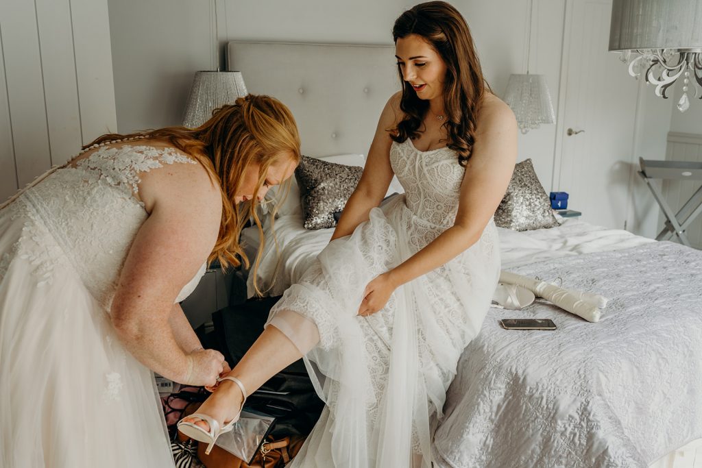 2 brides getting ready for their wedding in a silver grey bedroom helping put on their shoes