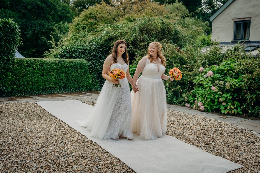 2 brides walking along a cream aisle carpet outdoors surrounded by greenery to their wedding ceremony carrying orange flower bouquets