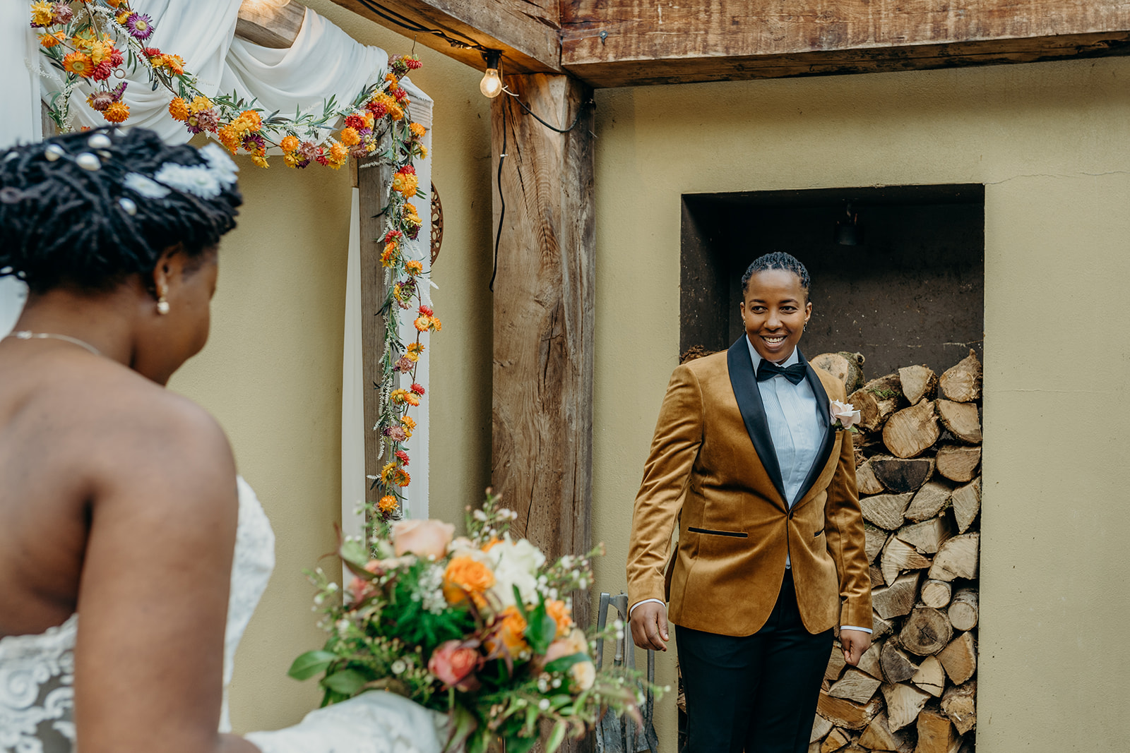 black same sex couple bride in a long white wedding gown walking into ceremony carrying orange and white bouquet other person dressed in gold velvet jacket and black bow tie