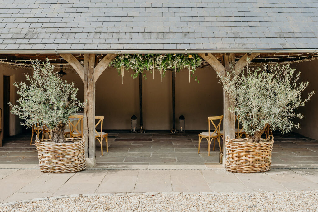 front on view of a oak framed wedding barn hung with a flower garland flanked by two large olive trees in baskets 