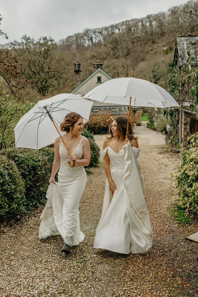 two brides in white wedding dresses holding white umbrellas walking outside under grey skies