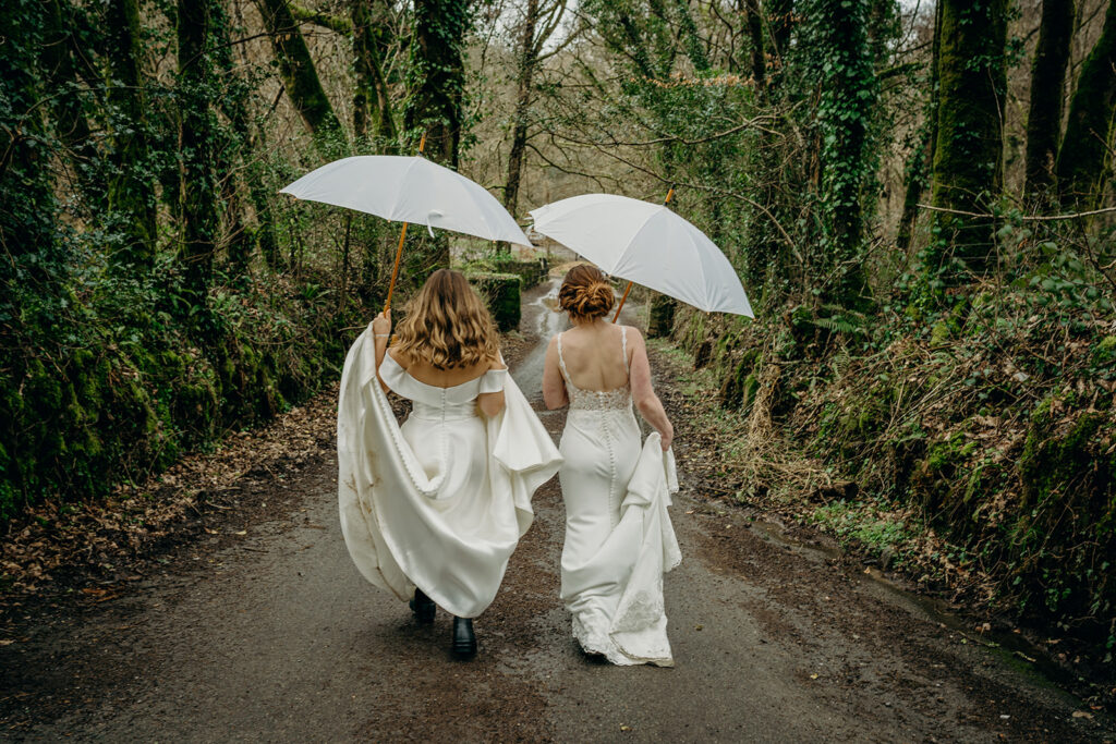 rear view of two brides in long white wedding dresses walking over a bridge in the woods holding their dresses and white umbrellas