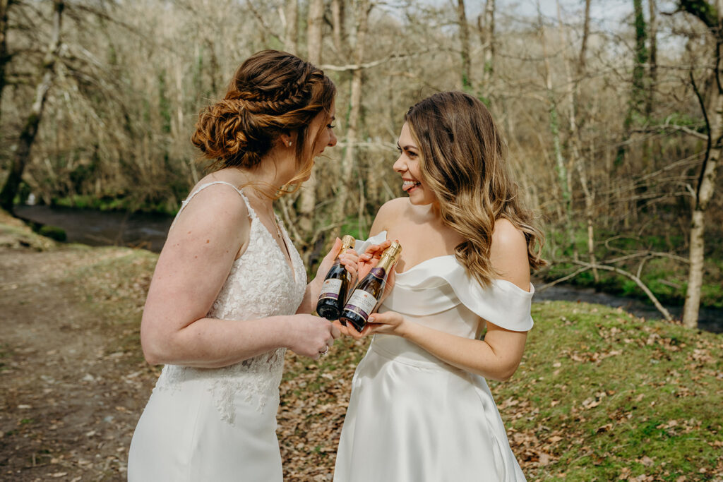 two brides outdoor in woodland holding mini prosecco bottles and laughing