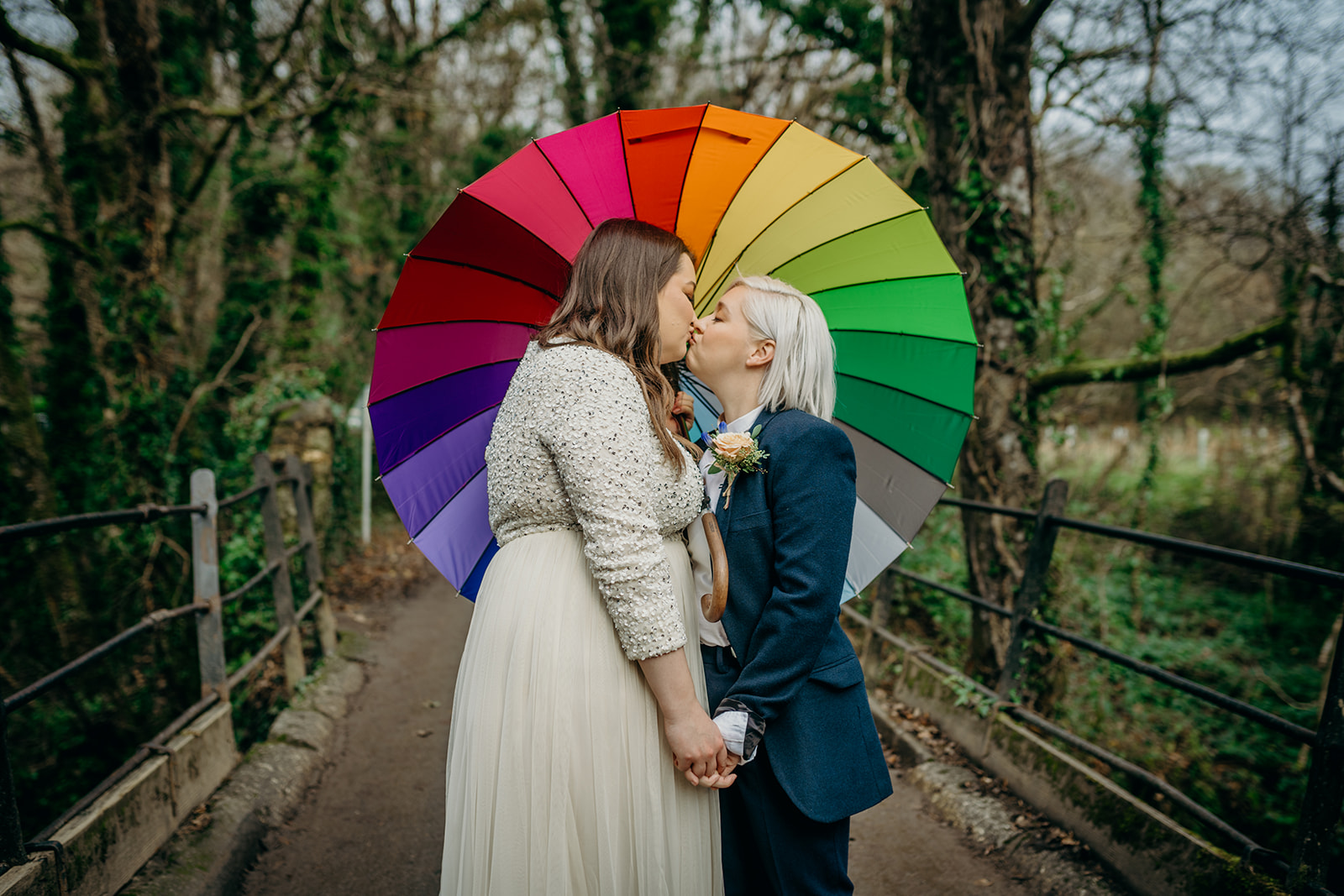 white same sex couple outside kissing on a rustic bridge one bride in a long white dress with long sleeves, the other bride in a dark blue trouser suit under a rainbow pride umbrella