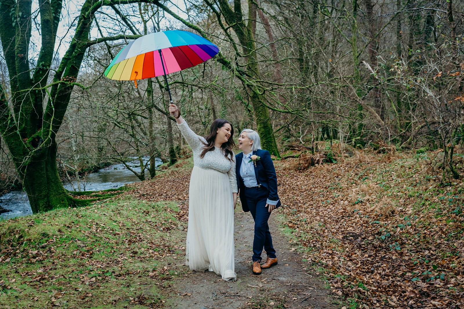 same sex couple two white brides in autumn woods one dark hair one blonde one in long white dress one in a dark blue trouser suit carrying rainbow pride umbrella aloft