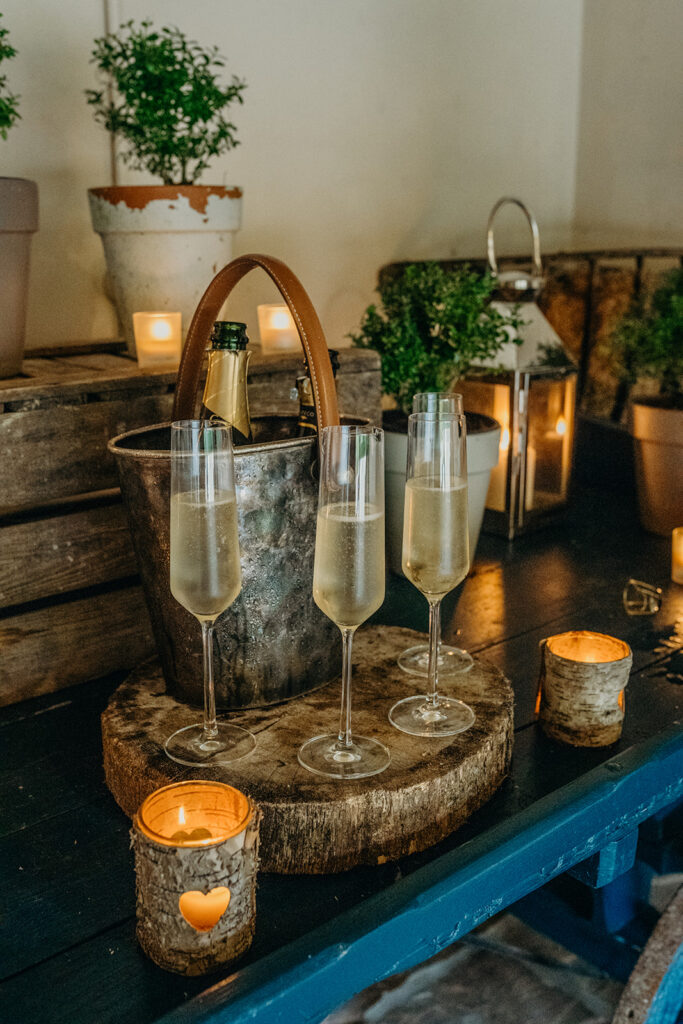 rustic dark blue painted handcart with tarnished metal wine bucket with leather handle containing open champagne bottle four full champagne flutes on a log slice with lit bark votives 