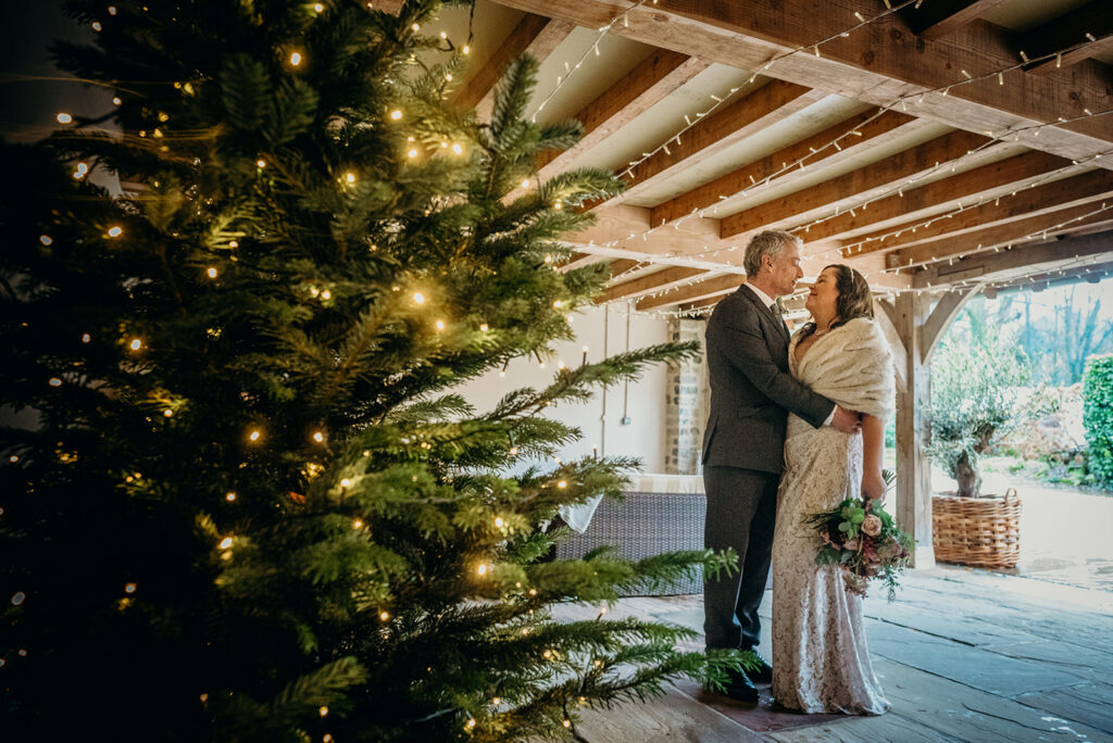 older bride and groom embracing in an oak wedding barn stood by christmas tree decorated with white fairy lights bride wearing fur wrap