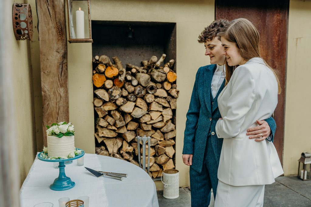 two brides in suits looking at a small white wedding cake on a blue cake stand with a log stack in the background
