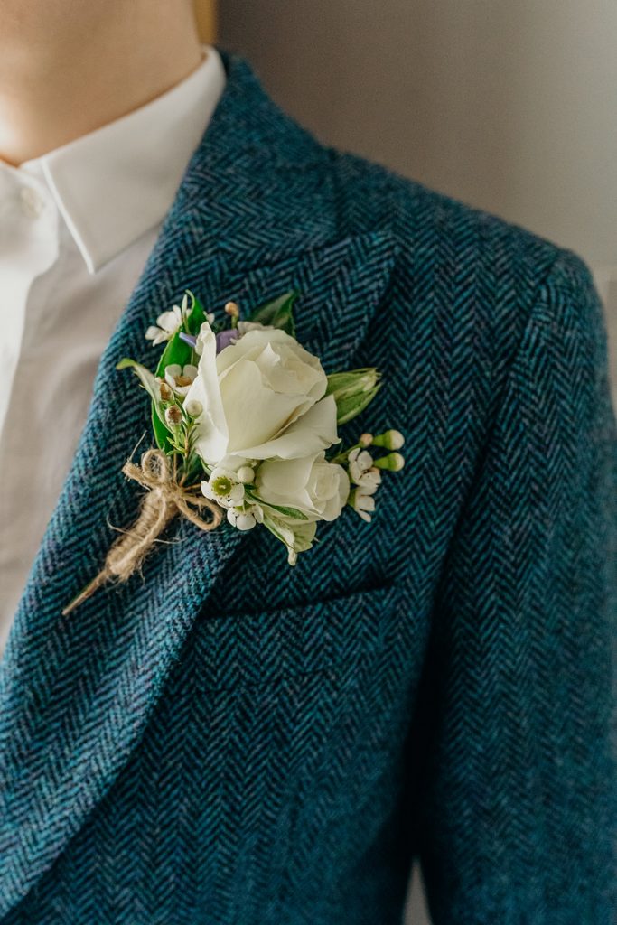 close up of a white boutonniere on a blue tweed suit jacket with white roses and was flowers tied with string