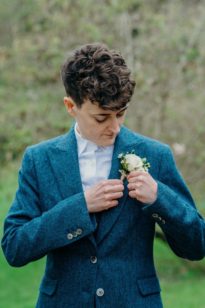 a bride with short hair in a blue tweed suit putting on a white rose boutonniere