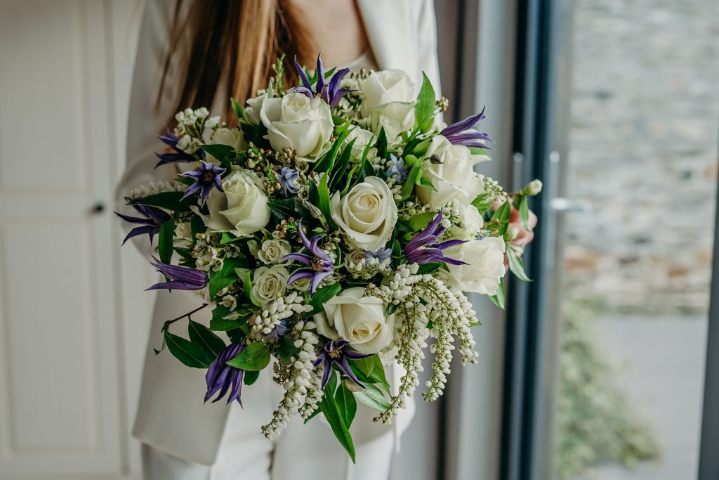 close up of a bride with long hair in a white trouser suit holding a white rose and blue clematis bouquet 