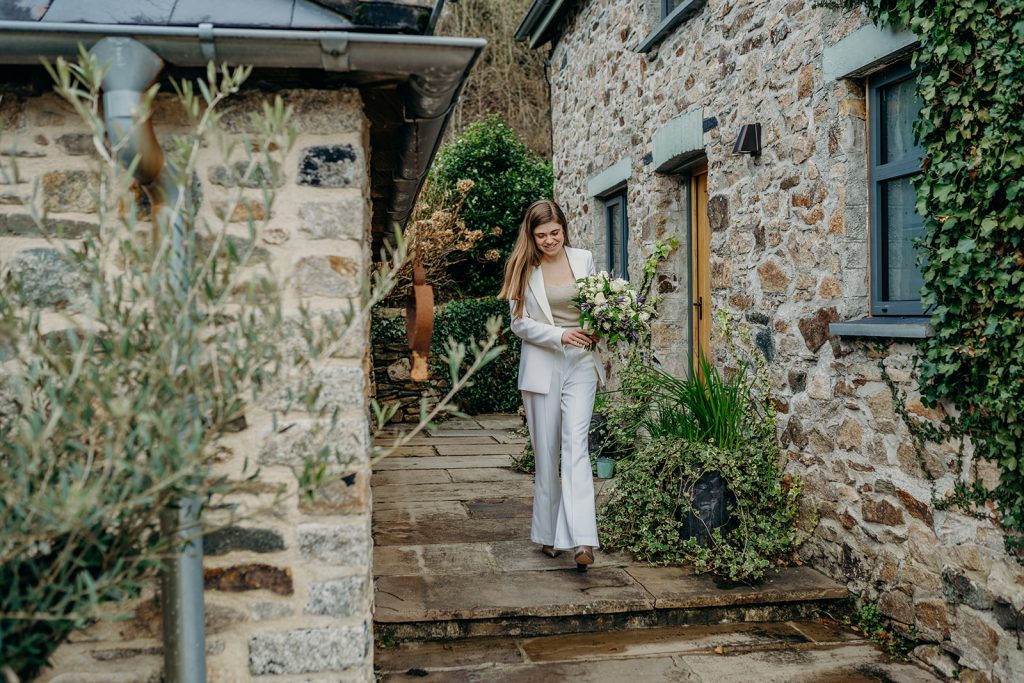 a bride in a white trouser suit carrying a white bouquet walking between 2 stone walls with foliage