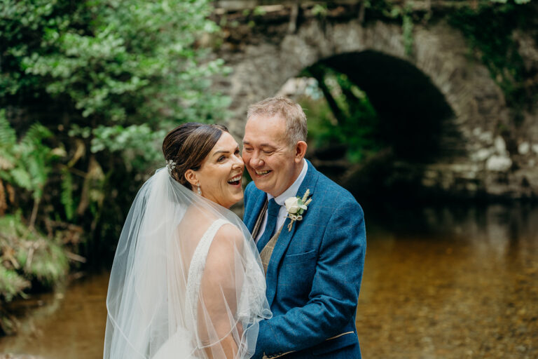 top half body shot of bride and groom laughing in front of stone arched bridge with river in background