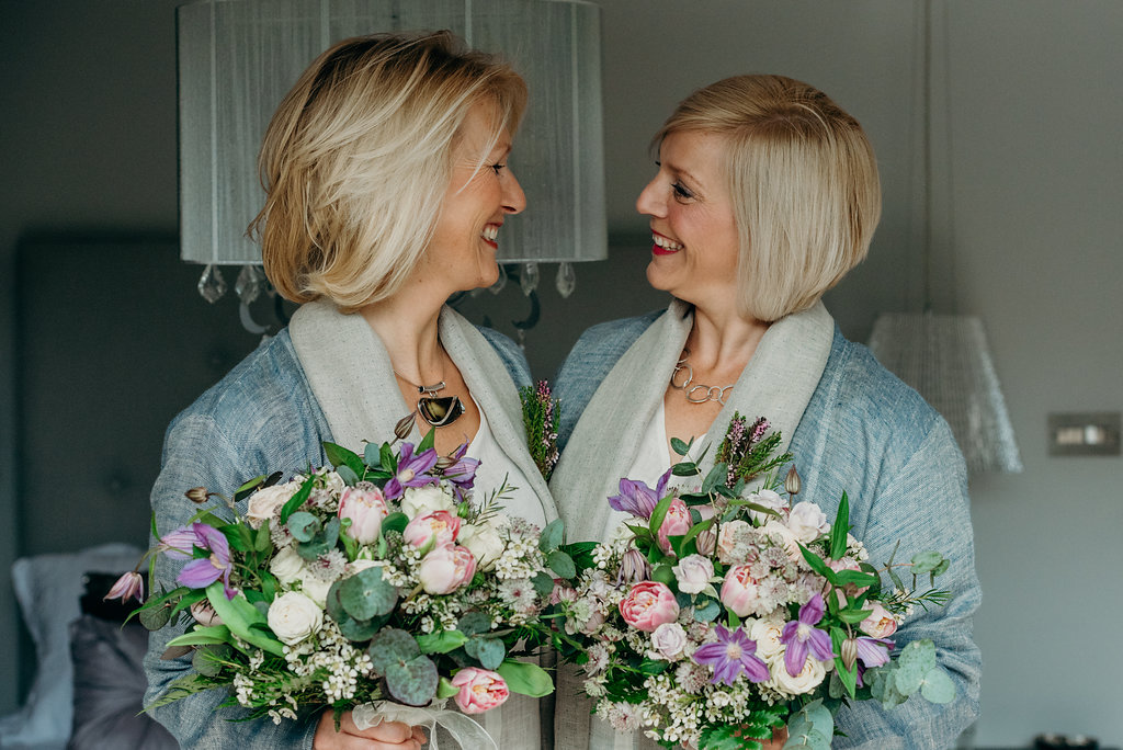 2 blond brides dressed in grey linen looking at each other holding pink and purple bouquets