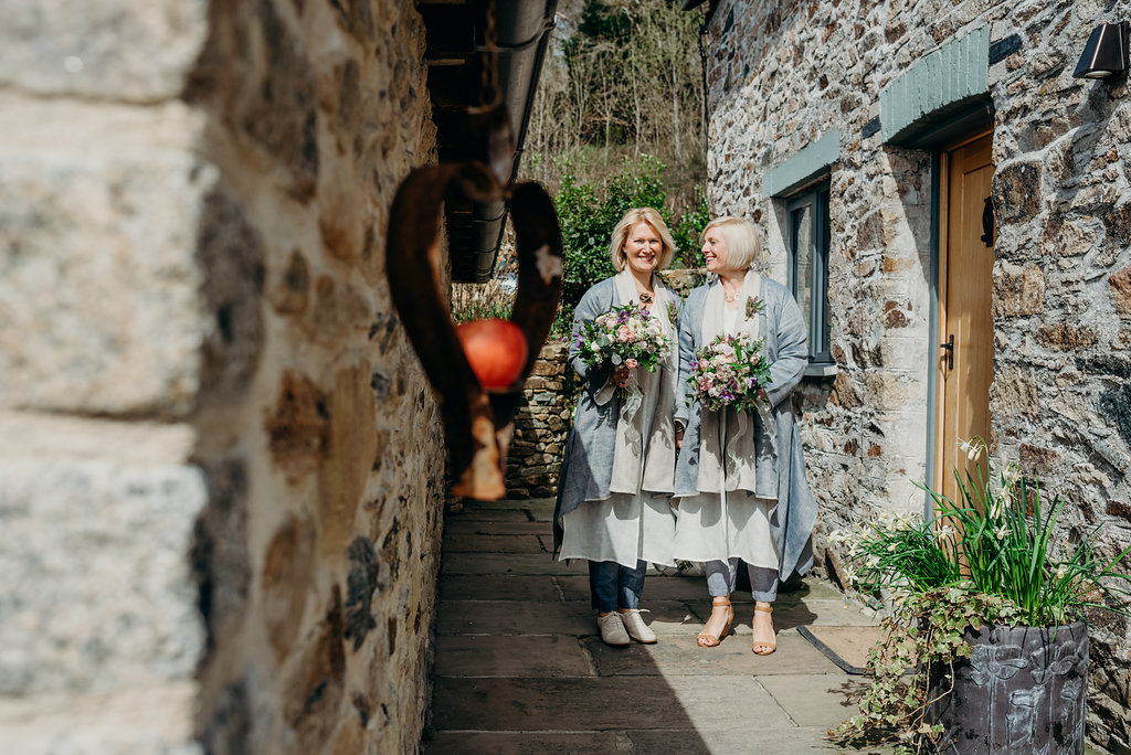two white blond brides dressing in matching grey linen flowing trouser suits holding bouquets outside a stone cottage before walking in to be married