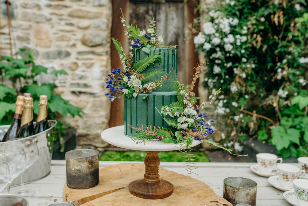 dark green iced cake decorated with fresh flowers on a marble and wood cake stand
